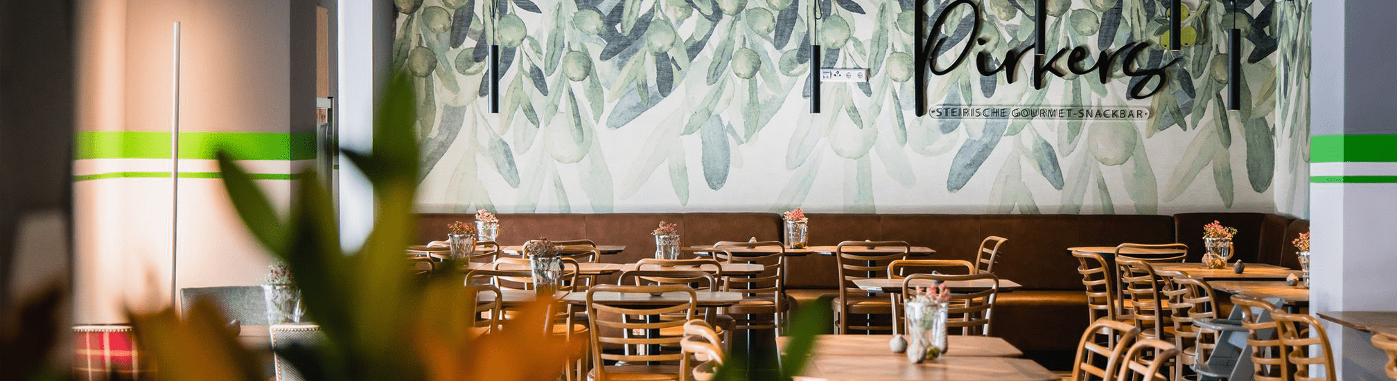 Pirkers steirische Gourmet-Snackbar: moderner Gastraum mit Oliven-Wandbild, Holzstühlen und Vasen.