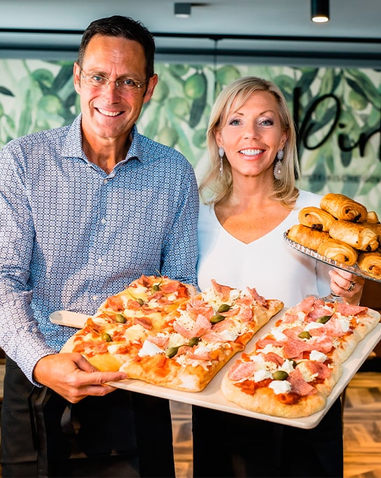Pirker Bistro: Frau Pirker und Herr Rippel präsentieren Blechpizza auf Holzbrettern und ein Tablett frischer Croissants.