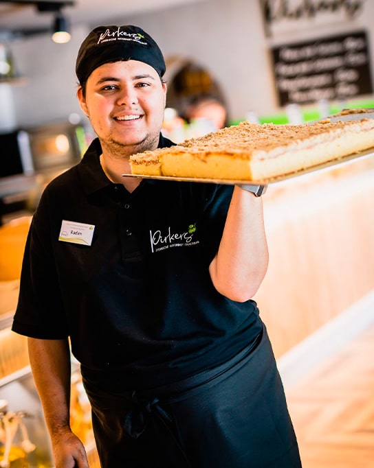 Pirker Bistro: Mitarbeiter im schwarzen Outfit trägt lächelnd ein großes Blechkuchen aus der Backstube.