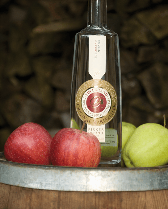 Pirker Edelbrand aus Apfel – Flasche auf Holzfass mit roten und grünen Äpfeln, Produktfoto.