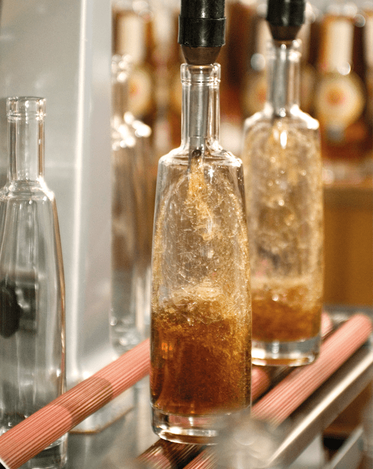 Abfüllanlage: Glasflaschen werden mit bernsteinfarbener Flüssigkeit befüllt, sprudelnde Nahaufnahme.