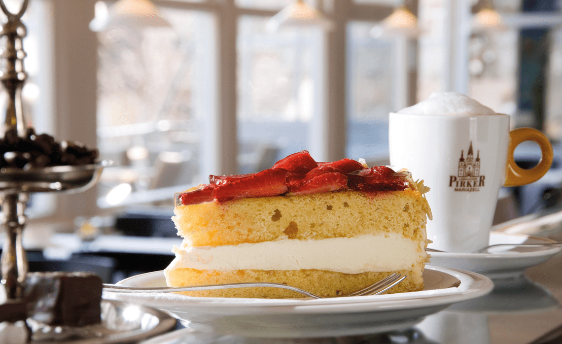 Pirker Café Mariazell: Erdbeerbiskuit mit Oberscreme auf Teller, Cappuccino im Hintergrund.