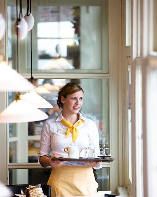 Café Mariazellerhof: Servicekraft mit Tablett und Kaffeeservice am Fenster, gemütliches Interieur.