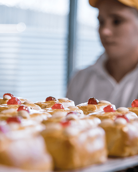 Pirker Backstube: Blech mit glasierten Küchlein und kandierten Kirschen, Mitarbeitende im Hintergrund.