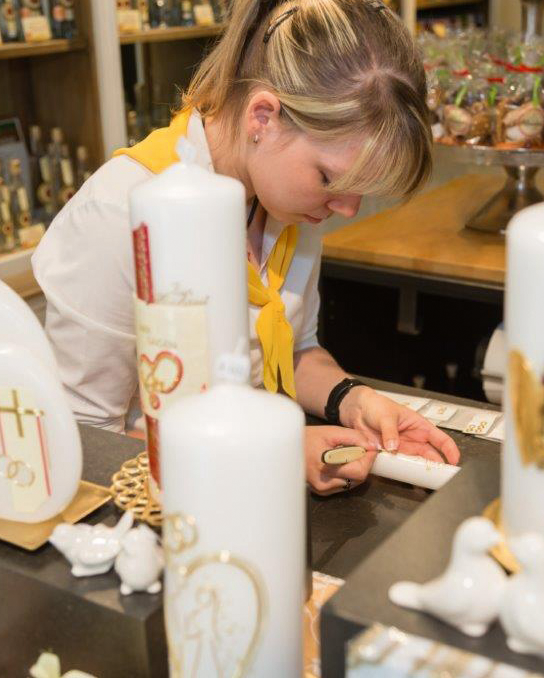 Pirker Shop: Mitarbeiterin verziert eine Kerze von Hand; weiße Festkerzen und Geschenke auf dem Tisch.
