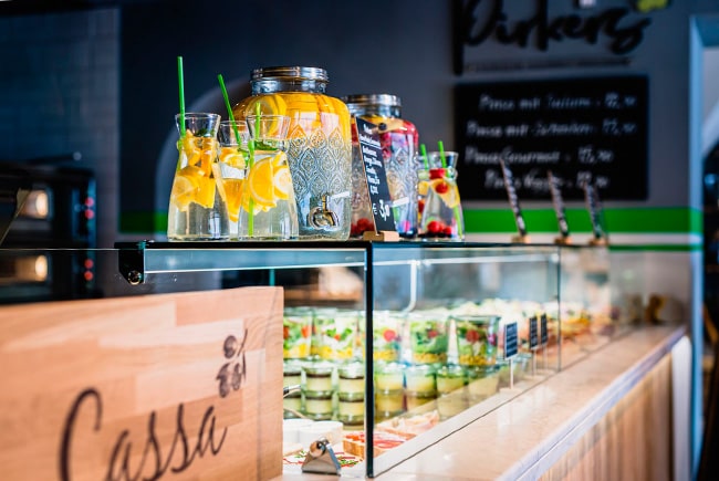 Pirker Bistro Theke: Vitrine mit Salaten, vorn Dispenser mit Zitronen- und Beeren-Limonade, „Kassa“-Schild.