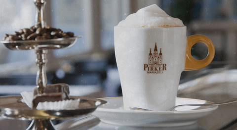 Pirker Café: Cappuccino mit Milchschaum in Tasse mit Logo, Löffel auf Untertasse, Dessertetagere daneben.