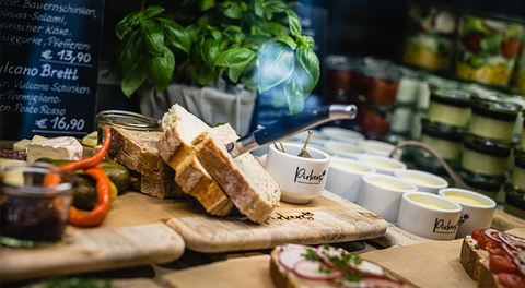 Pirker Bistro Vitrine: frisches Brot, Aufstriche und Antipasti; Basilikum, Jausen- & Salatgläser.