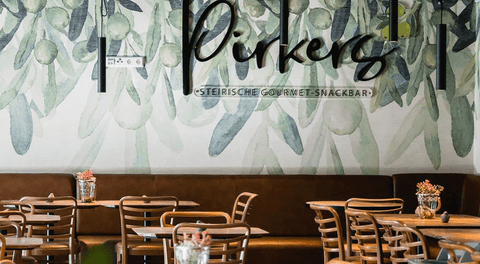 Pirkers steirische Gourmet-Snackbar: heller Gastraum mit Oliven-Wandbild, Holzstühlen und Vasen.