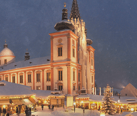 Mariazell im Winter: beleuchtete Basilika und Adventmarkt im Schnee, stimmungsvolle Abendszene.